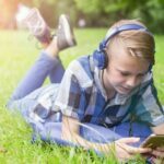 Laughing_kid_boy_12-16_year_old_with_headphones_outdoors._Childhood._Schoolboy._Lying_on_grass,_summer.